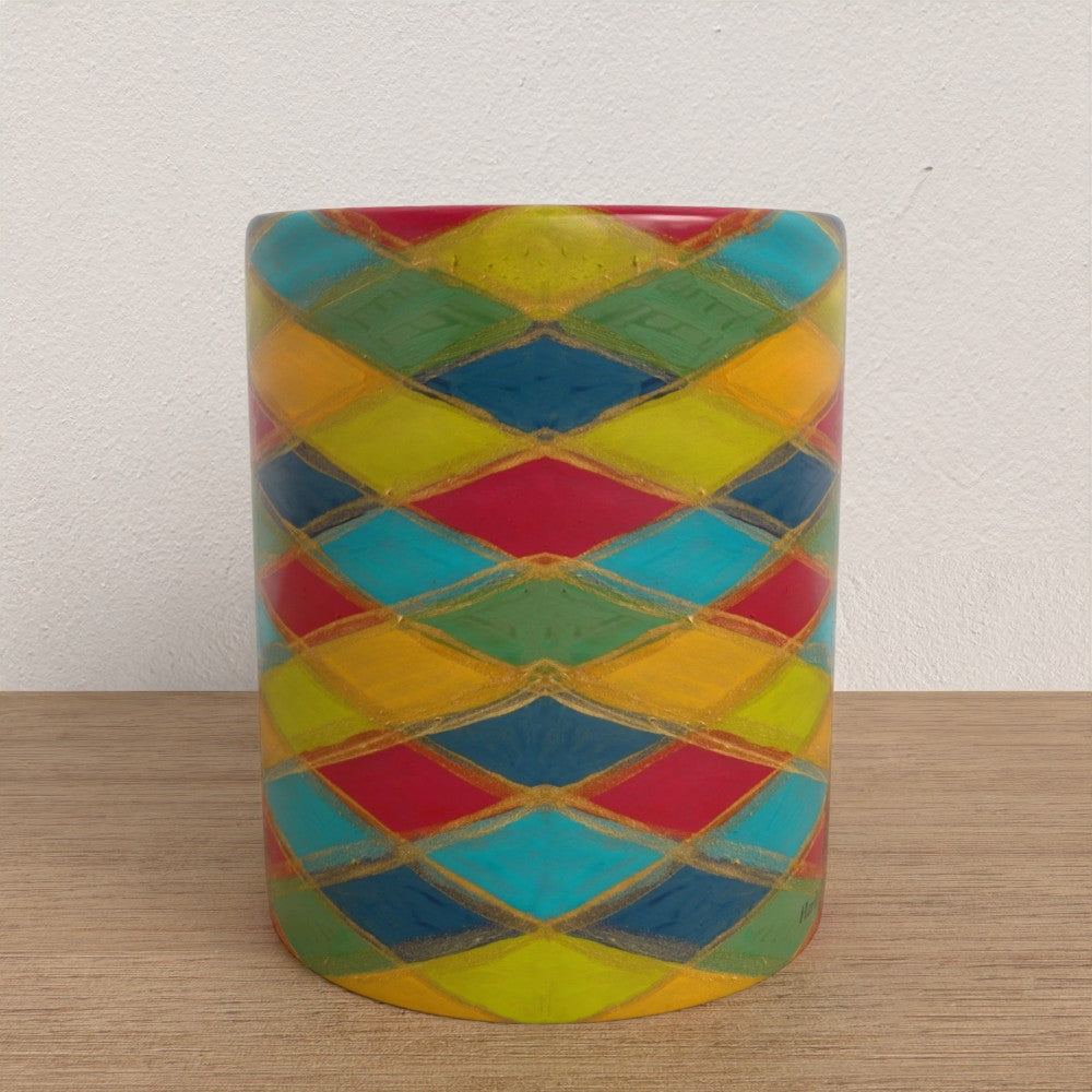 A mug with a colourful harlequin diamond pattern in red, yellow, green, and blue, separated by gold metallic lines, set on a wooden surface against a white wall.