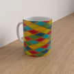 A white ceramic mug with a colourful geometric harlequin diamond pattern in yellow, red, green, and blue, sitting on a light wood tabletop against a plain off-white wall.
