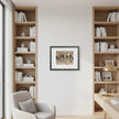 A framed art print featuring black ink figures on a beige background, displayed in a black frame on a white wall between two tall wooden bookshelves in a modern home office setting.