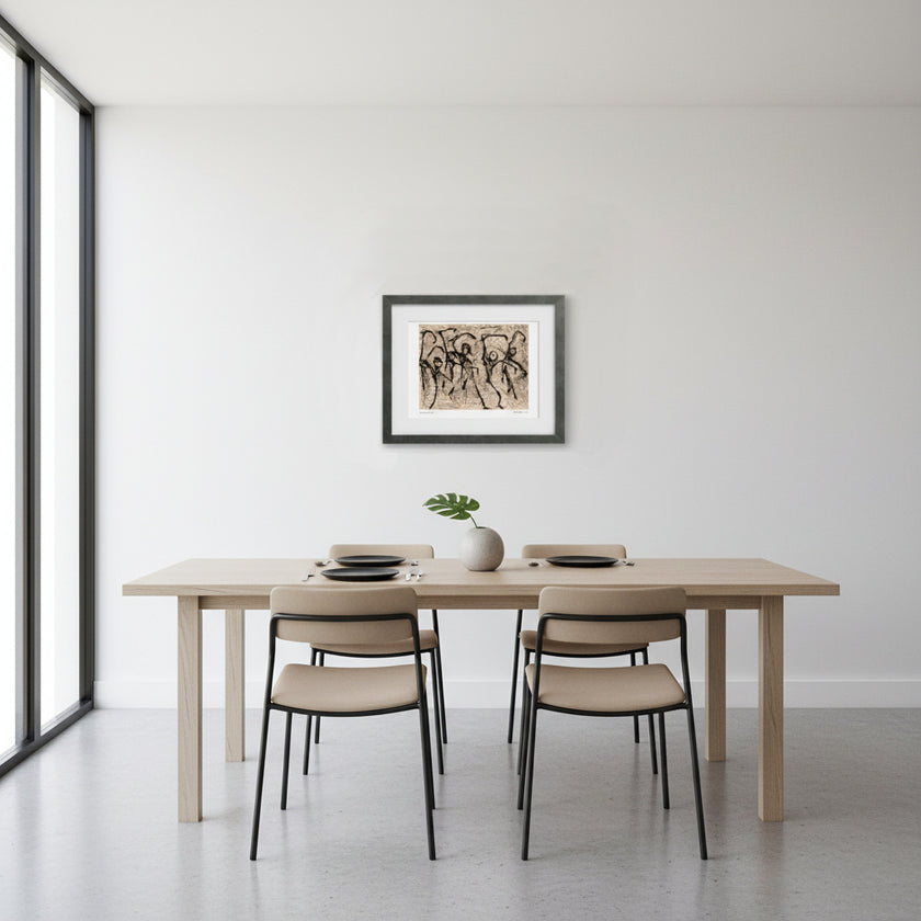 A minimalist dining room scene featuring a light wood rectangular table with four beige upholstered chairs, set against a white wall with a framed abstract print hanging above. A small white vase with a green plant sits on the table.