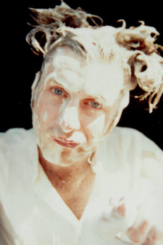 John Blake with messy light hair and a white shirt against a solid black background covered in foam