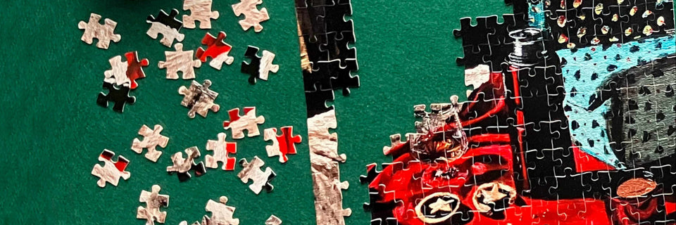 A close-up of a jigsaw puzzle being assembled on a dark green surface, with many loose puzzle pieces scattered on the left and a partially completed section on the right showing red, black, white, and blue details.