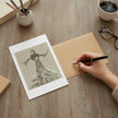 A hand holds a black fountain pen over a kraft paper envelope on a wooden desk, next to a printed art card featuring a black and white drawing of Ginger Rogers. The scene includes a cup of coffee, eyeglasses, pencils, and a book, creating a cosy, creative workspace atmosphere.