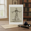 A greeting card with a black and white drawing of Ginger rogers, displayed on a wooden table with an envelope, fountain pen, and ink bottle in a softly blurred indoor setting.