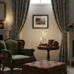 A styled vintage living room scene featuring a green velvet wing-back armchair, a dusty rose tufted ottoman, patterned grey curtains, a round wooden side table with candles and books, and a framed black-and-white print on the wall.