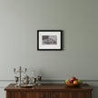 A framed black and white still life art print hangs on a light gray wall above a dark wood sideboard with silver goblets, a glass decanter, and a bowl of fruit.
