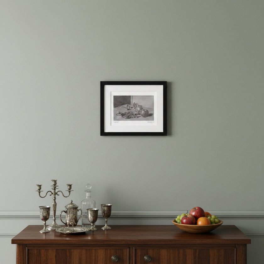 A framed black and white still life art print hangs on a light gray wall above a dark wood sideboard with silver goblets, a glass decanter, and a bowl of fruit.