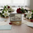 A greetings card showing a drawing of flowers and fruit on a table with lilies and roses and cards