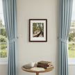 A framed art print depicting a sunlit forest path hangs on a beige wall between two light blue curtains. A small round wooden table below holds a mug, papers, and stacked books.