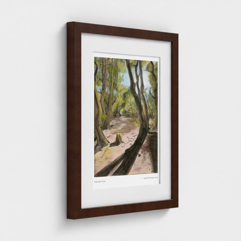 A framed art print depicting a forest path with tall trees, shown at a slight angle against a light grey wall. The print features a white mat border and is housed in a dark brown wooden frame.