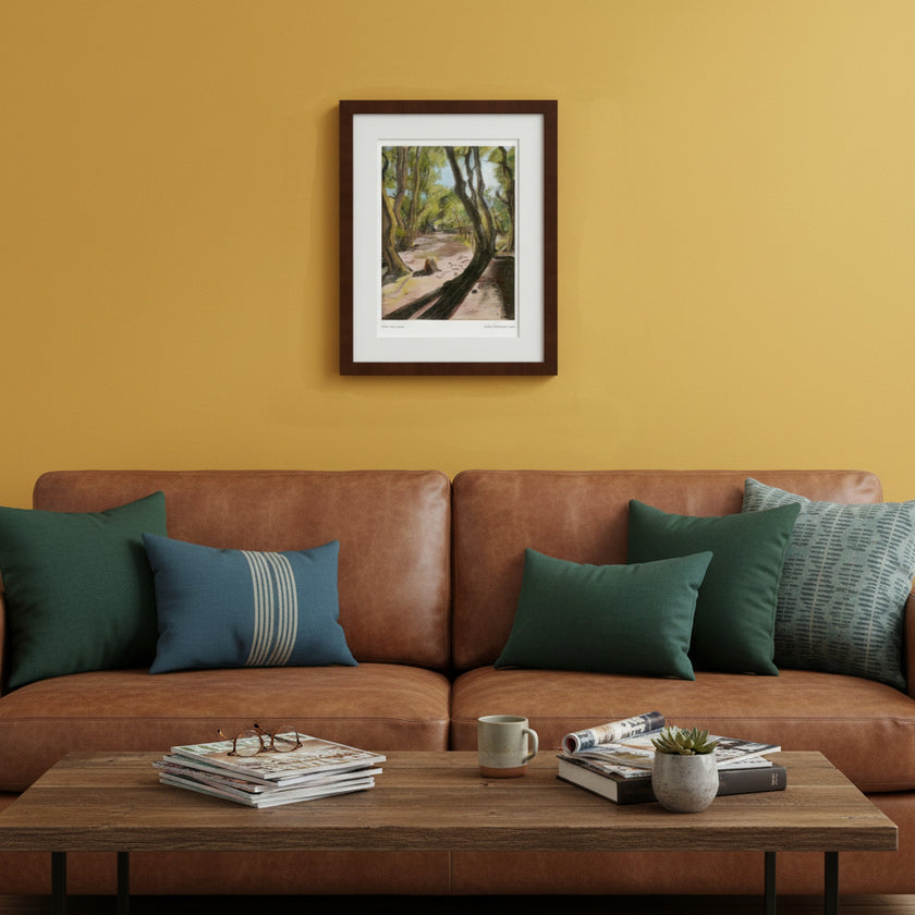 A framed art print depicting a forest path hangs on a mustard yellow wall above a brown leather sofa with green and blue throw pillows. A wooden coffee table with books and a mug sits in the foreground.