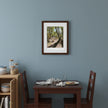 A framed art print depicting a sunlit forest path with trees and dappled light, hung on a dusty blue-gray wall above a wooden dining table and chairs.