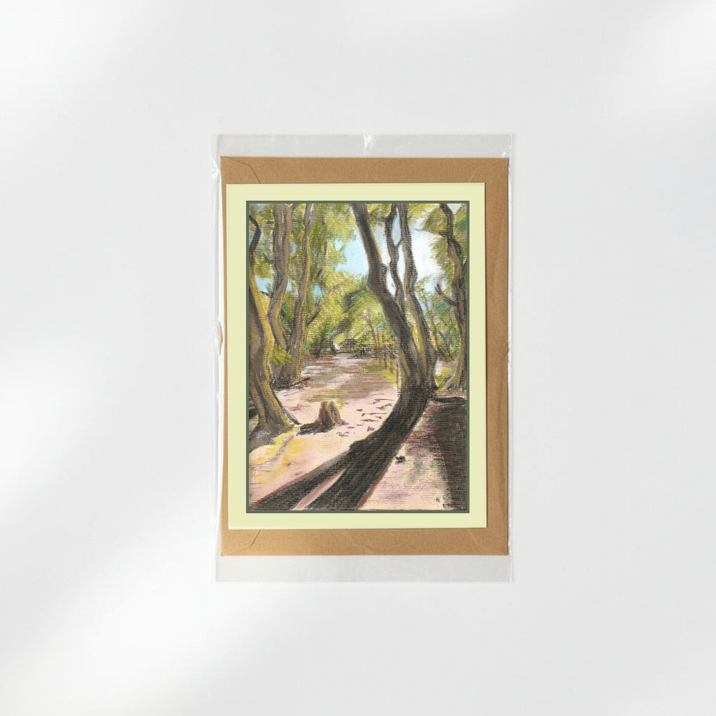 A greeting card featuring a painted forest path scene, presented in a clear sleeve with a kraft envelope behind it on a white background.