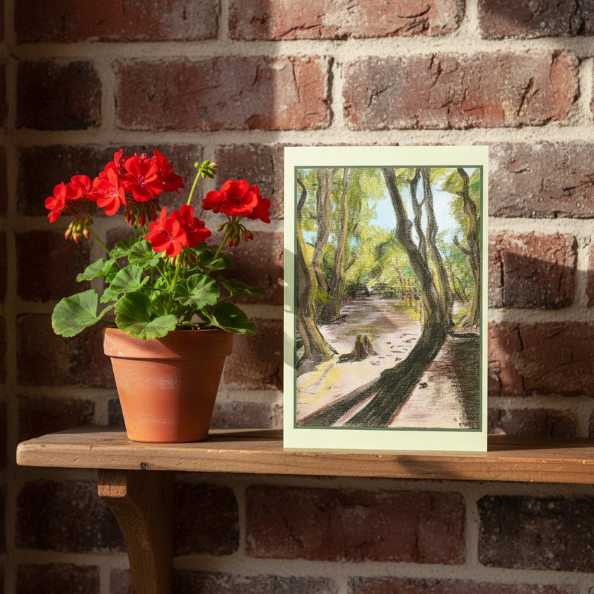 A greetings card depicting a sunlit forest path with tall trees, styled on a wooden shelf next to a potted red geranium against a red brick wall.