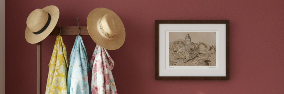 A framed art print on a red wall next to a coat stand with shirts and hats