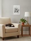 Edith the Cat Art Print on a wall above an armchair with a sleeping cat and sidetable with plant and lamp
