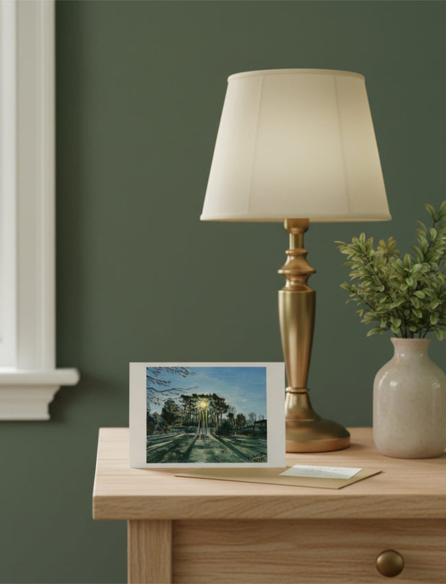 Winter Sun Through Trees Greetings Card on a table with a lamp and foliage vase