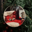 A round ceramic Christmas ornament hanging from a gold loop, featuring a printed festive scene with red fabric, white fur, a dark bottle, and holiday treats, displayed against green artificial pine branches.