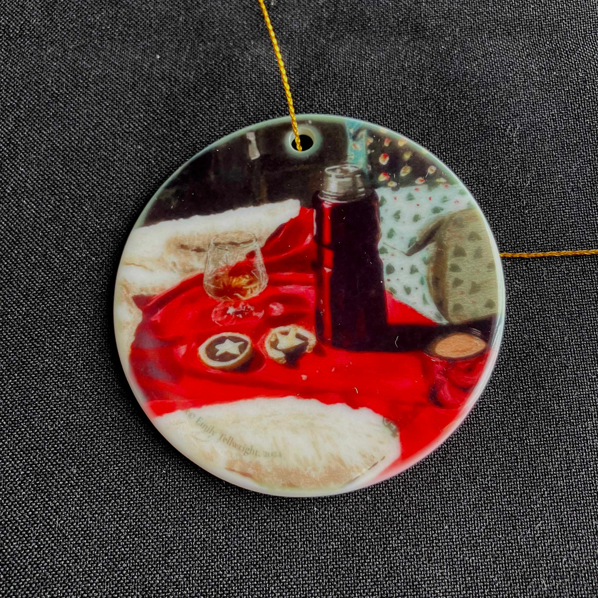 A round ceramic ornament featuring a printed still life scene with a red cloth, a dark bottle, a glass, and white fluffy shapes, hanging from a thin gold string against a dark textured fabric background.