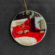 A round ceramic ornament featuring a printed still life scene with a red cloth, a dark bottle, a glass, and white fluffy shapes, hanging from a thin gold string against a dark textured fabric background.