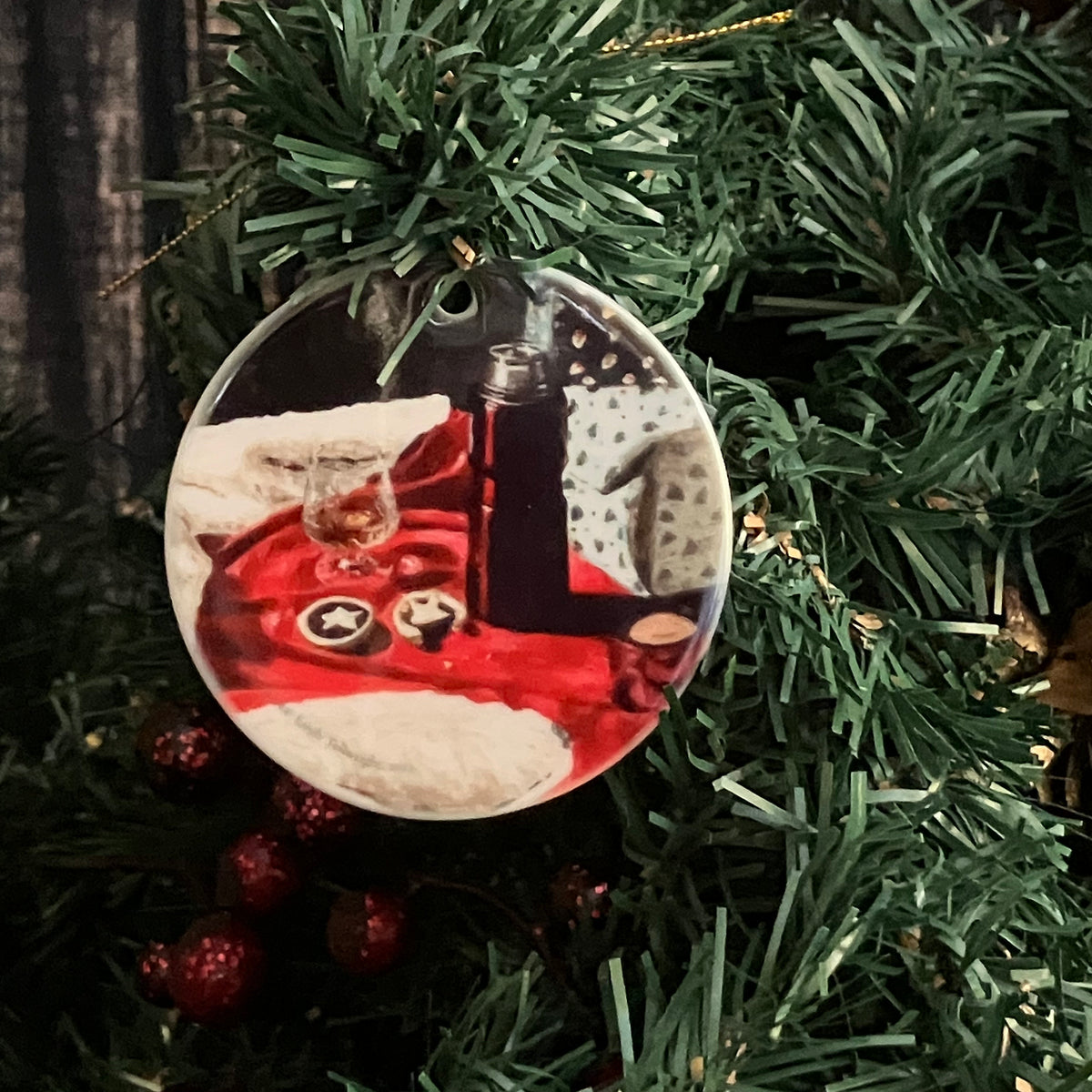 A round ceramic Christmas ornament hanging from a gold loop, featuring a printed festive scene with red fabric, white fur, a dark bottle, and holiday treats, displayed against green artificial pine branches.