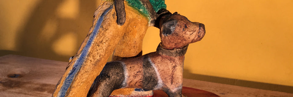 A rustic hand-paintedfigurine depicting a dog standing on its hind legs and holding a yellow-and-blue staff, set against a warm yellow background.