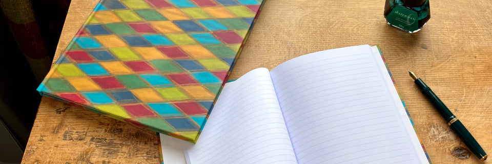 A colorful diamond-patterned notebook cover lies next to an open lined notebook on a wooden desk, with a green ink bottle and fountain pen nearby.