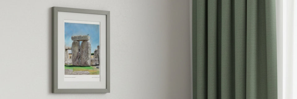 a framed print of a drawing of stonehenge on a cream wall next to green curtains