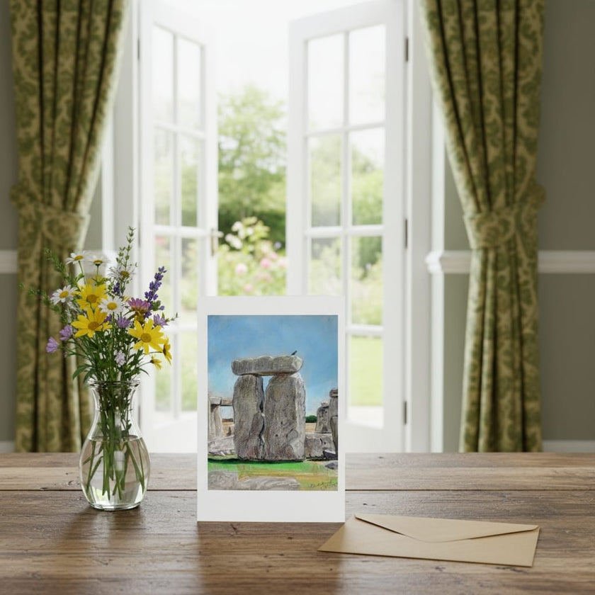 A greetings card of Stonehenge displayed on a wooden table next to a glass vase of yellow and purple wildflowers, with a brown envelope in front. The print is set against a real-world background of a bright room with green curtains and open white French doors.