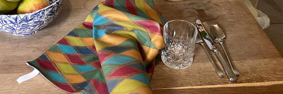 A colorful geometric patterned cloth napkin or table linen spread on a wooden table next to a clear cut-glass tumbler, silver cutlery, and a blue-and-white fruit bowl.