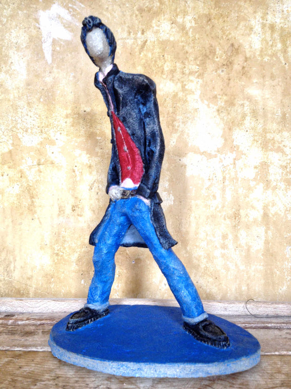 A stylised figurine of a walking teddy boy wearing a black coat, blue trousers, and a red shirt, standing on a round blue base against a textured beige wall.