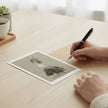 A hand holds a black pen over a greetings card showing ginger rogers on a light wood desk, with a cup and small plant in the softly blurred background.