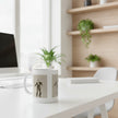 A white ceramic mug with a sepia-toned graphic print of a dancer, sitting on a white desk in a bright, modern home office setting.
