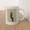 A white ceramic mug with a printed sepia-toned illustration of a dancing figure, placed on a light wood tabletop against a white wall.