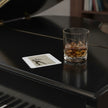 A clear square coaster with rounded corners sits on the glossy black surface of a grand piano next to a glass of whiskey with ice.