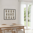 A framed black and white art print featuring twelve minimalist human figure sketches hangs on a white wall above a light wood dining table and chairs in a bright, modern room with white curtains and a glass door overlooking a green garden.