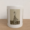 A white ceramic mug with a sepia-toned vintage illustration of a woman in a patterned dress, standing on a light wood surface against a textured off-white wall.