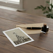 A printed card with a black-and-white illustration of Ginger Rogers lies on a wooden table beside a brown envelope, a black fountain pen, and a small glass ink bottle.
