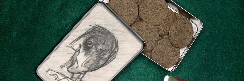 A square metal tin with a sketch of a face on the lid, shown next to an open tin filled with round brown cookies on a dark green textured background.