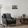 A black and white landscape art print of a tree is displayed on a white wall in a styled living room setting with a dark gray leather armchair, a wooden side table, a potted plant, and a floor lamp.