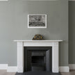 A framed black and white landscape sketch hangs on a sage green wall above a white fireplace mantel with a small gold-toned clock on it.