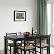 A black wooden dining table with four matching chairs featuring natural woven seats, styled with a wine bottle, two glasses of red wine, and a fruit bowl on a white wall in a bright room with green curtains.