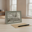A monochrome sketch of an outdoor woodland scene greetings card displayed on a light wood table, with a kraft envelope and black pen in front and a softly blurred interior background.