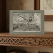 A monochrome landscape sketch of a tree in a forest greetings card, displayed on a carved wooden mantel with a Celtic knot and dragon design.