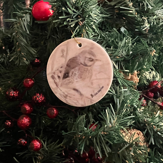 A round ceramic Christmas ornament featuring a detailed sketch of a bird, hanging from a gold loop on a green Christmas tree with red ornaments.