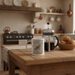 A white ceramic mug with a detailed bird illustration sits on a rustic wooden table in a cosy, warm-toned kitchen setting, alongside a French press and a basket of pastries.