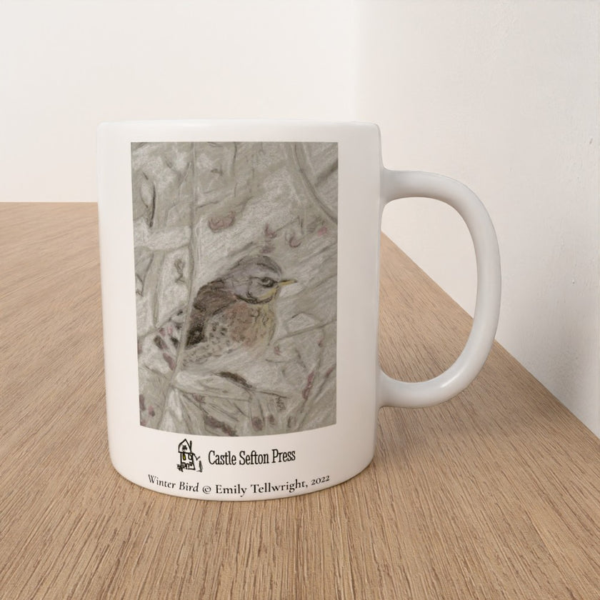 A white ceramic mug with a printed winter bird illustration and the text 'Castle Sefton Press' and 'Winter Bird by Emily Tellwright, 2022', sitting on a light wood surface against a white wall.