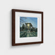 A framed art print depicting a forest path with tall trees, shown at a slight angle against a light grey wall. The print features a white mat border and is housed in a dark brown wooden frame.