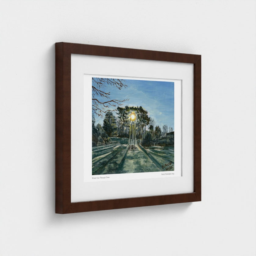 A framed art print depicting a forest path with tall trees, shown at a slight angle against a light grey wall. The print features a white mat border and is housed in a dark brown wooden frame.