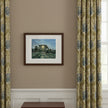 A framed art print depicting a sunlit forest path hangs on a beige wall between two light blue curtains. A small round wooden table below holds a mug, papers, and stacked books.